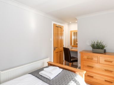 A bedroom with a wooden bed frame white folded towels on the bed a wooden dresser with a plant and a small desk with a chair and mirror at Spring Cottage in Ambleside