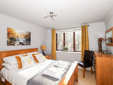 A bedroom with a wooden bed frame bedding and pillows a window with yellow curtains a wooden dresser with a chair and wall art at Spring Cottage in Ambleside