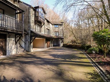 A driveway leading to garages of stone houses with trees and bushes nearby at Spring Cottage in Ambleside
