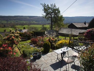 A garden with flowering bushes and a table with chairs on a stone patio overlooking fields and hills at Dormouse Cottage in Coniston