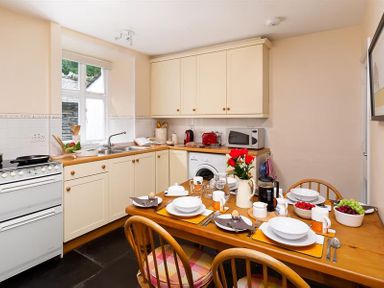 A kitchen with cream cabinets a wooden dining table set for breakfast with chairs at Dormouse Cottage in Coniston