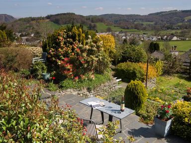 A garden with a table and chairs surrounded by bushes and flowers with hills and houses in the background at Dormouse Cottage in Coniston