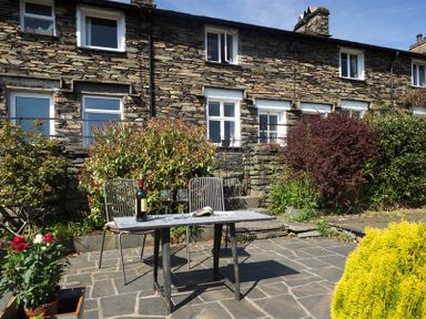 A stone patio with a table and two chairs a bottle of wine and two glasses in front of a stone house with shrubs at Dormouse Cottage in Coniston