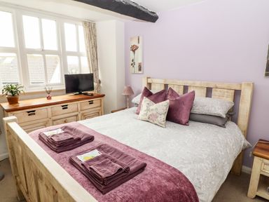 A bedroom with a double bed with pillows and folded towels on a purple throw a wooden dresser with a television and a plant by the window at Buttonhole Cottage Ambleside
