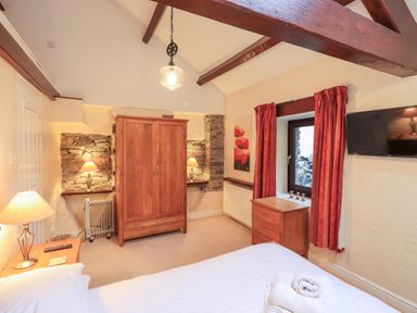 A bedroom with a bed a wooden wardrobe bedside tables lamps a window with red curtains and a wall-mounted TV at Old Coach House Ambleside