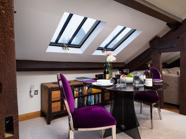 A dining area with a round black table set with dishes glasses and wine bottles two purple chairs under skylights and wooden beams at Hayloft in Windermere
