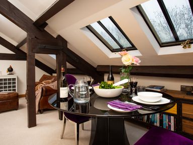 A dining area with a round glass table set for two with wine bottles and a bowl of salad under skylight windows at Hayloft in Windermere