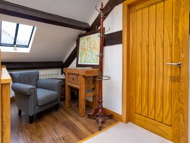 A small seating area with a grey armchair wooden desk and coat stand near a wooden door under a skylight window at Hayloft in Windermere
