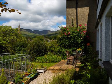 An outdoor garden area with a metal table and chairs surrounded by plants and flowers with mountains and a church steeple in the background at Uldale in Ambleside