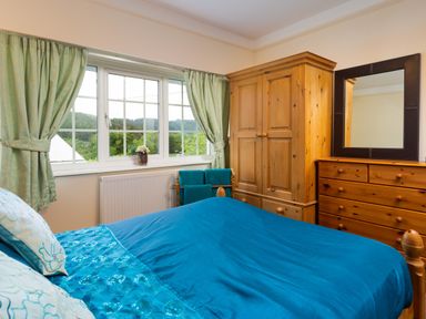 A bedroom with a bed with blue bedding a wooden wardrobe a wooden dresser with mirror and a window with green curtains at Uldale in Ambleside