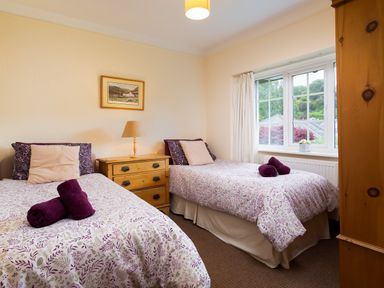 A bedroom with two single beds a wooden dresser with a lamp a window with curtains and a wardrobe at Uldale in Ambleside