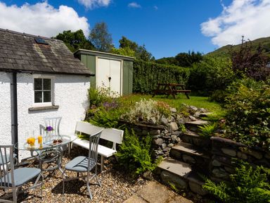 An outdoor garden area with a glass table and chairs next to a white house and stone steps leading to a grassy area with a wooden picnic bench at Uldale in Ambleside