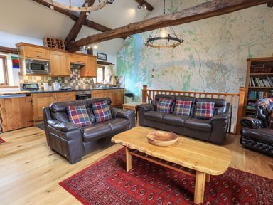 A living room with leather sofas and plaid pillows a wooden coffee table a red rug and a kitchen area with wooden cabinets at Tullythwaite Garth in Crosthwaite