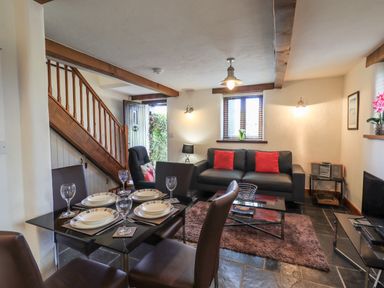A living and dining area with a black sofa with red cushions a glass dining table with place settings wooden stairs and a television at Bobbin Cottage in Ings