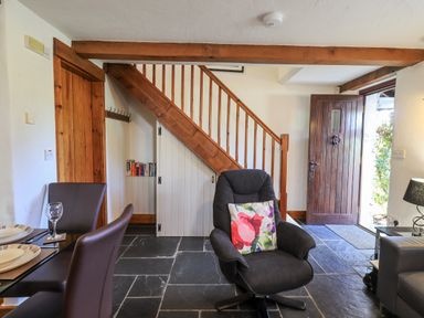 A room with a wooden staircase a dark armchair with a floral cushion dining chairs a wooden door and slate flooring at Bobbin Cottage in Ings