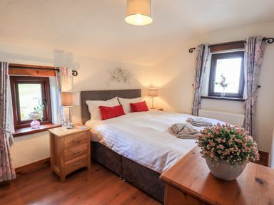 A bedroom with a bed with red cushions a plant on a wooden dresser two bedside tables with lamps and windows with floral curtains at Bobbin Cottage in Ings