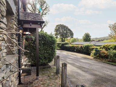 A stone exterior with a small porch and hanging basket next to a paved driveway with hedges and trees at Bobbin Cottage in Ings