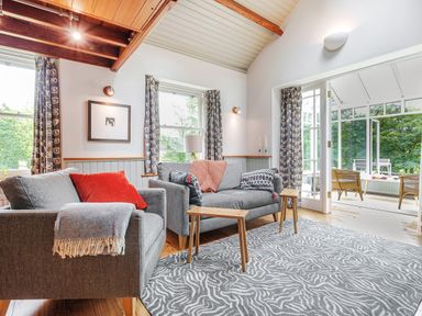 A sitting room with two gray sofas wooden side tables patterned curtains and an adjoining room with wicker chairs at The Stable At Oakbank Elterwater