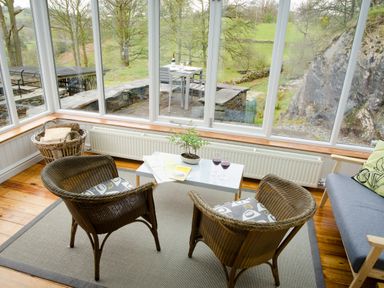 A sunroom with two wicker chairs a small white table with two glasses of red wine and a plant a basket of firewood and a sofa at The Stable At Oakbank in Elterwater