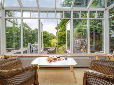 A conservatory with wicker chairs and a white table with fruit and bread outside greenery visible at The Stable At Oakbank in Elterwater
