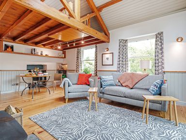 A living room with gray sofas, patterned rug, wooden beams and a dining table at The Stable At Oakbank in Elterwater