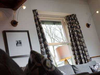 A window with patterned curtains and table lamp behind a sofa with cushions and wall lights at The Stable At Oakbank in Elterwater