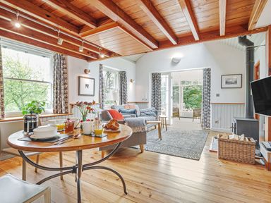 A dining table set with breakfast items near windows and a seating area with sofas a wood stove and a television in a living room at The Stable At Oakbank in Elterwater