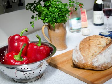 A kitchen counter with red bell peppers in a metal colander fresh bread on a wooden cutting board a brown jug with parsley and bottles in the background at The Stable At Oakbank in Elterwater