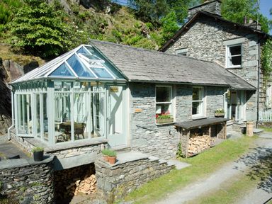 A stone house with a glass conservatory and stacked firewood outside at The Stable At Oakbank in Elterwater