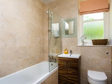 A bathroom with a bathtub shower combination a wooden cabinet with a sink a window with a small mirror and basket on the sill and a toilet at Highbank in Bowness