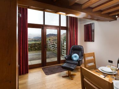 A living area with wooden dining chairs a black recliner chair with a blue cushion near glass doors with red curtains and a view of countryside hills at Nest Barn in Keswick
