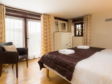 A bedroom with a brown bedspread two folded towels a black armchair a white chest of drawers and windows with curtains at Nest Barn in Keswick