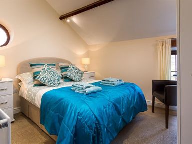 A bedroom with a bed covered with a blue bedspread and patterned pillows next to bedside tables with lamps and a chair near a window at Nest Barn in Keswick