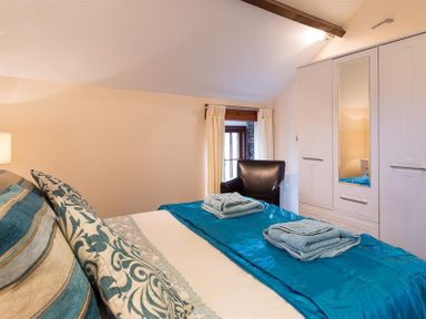 A bedroom with a bed with blue linens and folded towels a black armchair a window with curtains and a white wardrobe with a mirror at Nest Barn in Keswick
