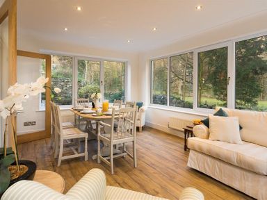 A dining area with a wooden table and white chairs next to large windows and cream sofas at Lanterns At Grasmere in Grasmere