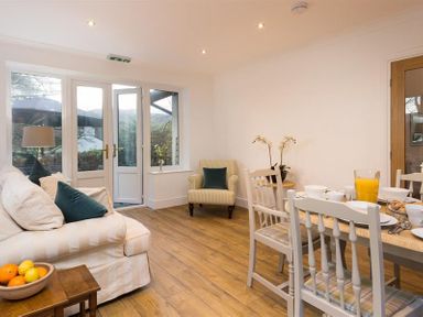A living area with a white sofa, armchair, wooden dining table set, and glass doors at Lanterns At Grasmere in Grasmere