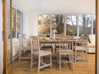 A dining area with a wooden table and six chairs near large windows and a sofa at Lanterns At Grasmere in Grasmere