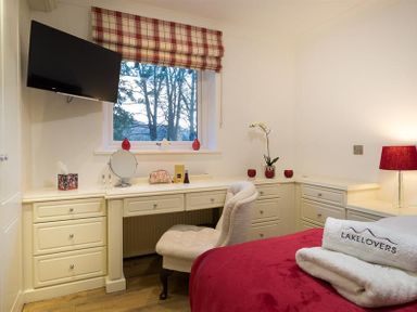 A bedroom with a red bedspread a white chair and built in cream drawers at Lanterns At Grasmere in Grasmere