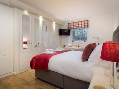 A bedroom with a bed red pillows and a red blanket a wall of white wardrobes with mirrors and a window with a red checkered blind at Lanterns At Grasmere in Grasmere