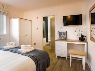 A bedroom with a bed, wardrobe, desk, stool, wall-mounted TV, and an open door leading to a bathroom at Lanterns At Grasmere in Grasmere