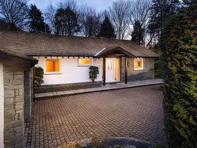 A house exterior with white walls a front door and lit windows surrounded by trees and bushes at Lanterns At Grasmere in Grasmere