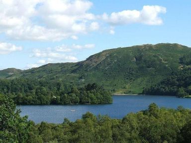 A lake surrounded by trees and hills under a blue sky with clouds at Lanterns At Grasmere in Grasmere