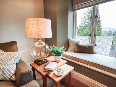 A corner with a window seat with cushions next to a sofa and wooden tables with a lamp plant books and a cup at Hart Head Barn in Rydal