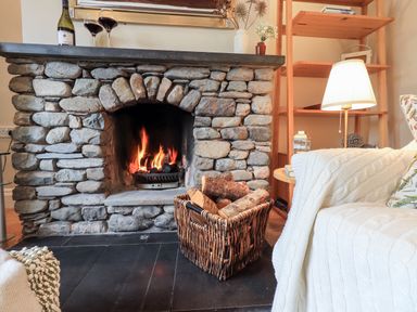 A stone fireplace with a fire burning and a basket of firewood next to a white knitted blanket on a sofa with a lamp and wooden shelving in the background at Hart Head Barn in Rydal