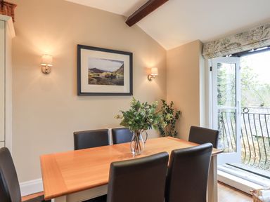 A dining area with a wooden table and six chairs a vase with flowers on the table wall lights a framed picture and a door opening to a balcony at Hart Head Barn in Rydal