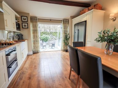 A kitchen with wooden floor cabinets a dining table with chairs and glass doors leading outside at Hart Head Barn in Rydal