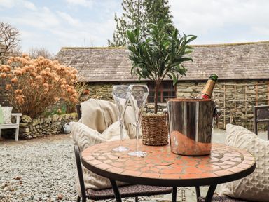 An outdoor table with two empty glasses a champagne bottle in an ice bucket and a small potted plant with pillows on chairs at Hart Head Barn in Rydal