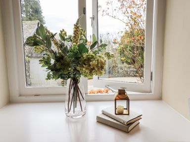 A vase with green flowers next to books and a candle holder on a white table by an open window at Hart Head Barn in Rydal