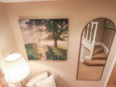 A corner of a room with a lamp chair and pillow under a tree artwork next to a mirror reflecting stairs at Hart Head Barn in Rydal