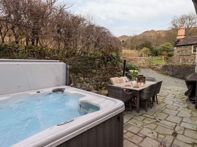 An outdoor patio area with a hot tub and a dining table with chairs set on a stone floor near a stone wall and countryside at Hart Head Barn in Rydal